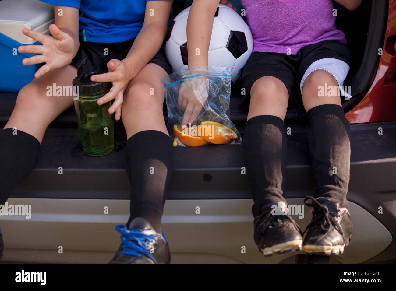 Waist down of boy and younger sister sitting in car boot eating oranges on football practice break Stock Photo