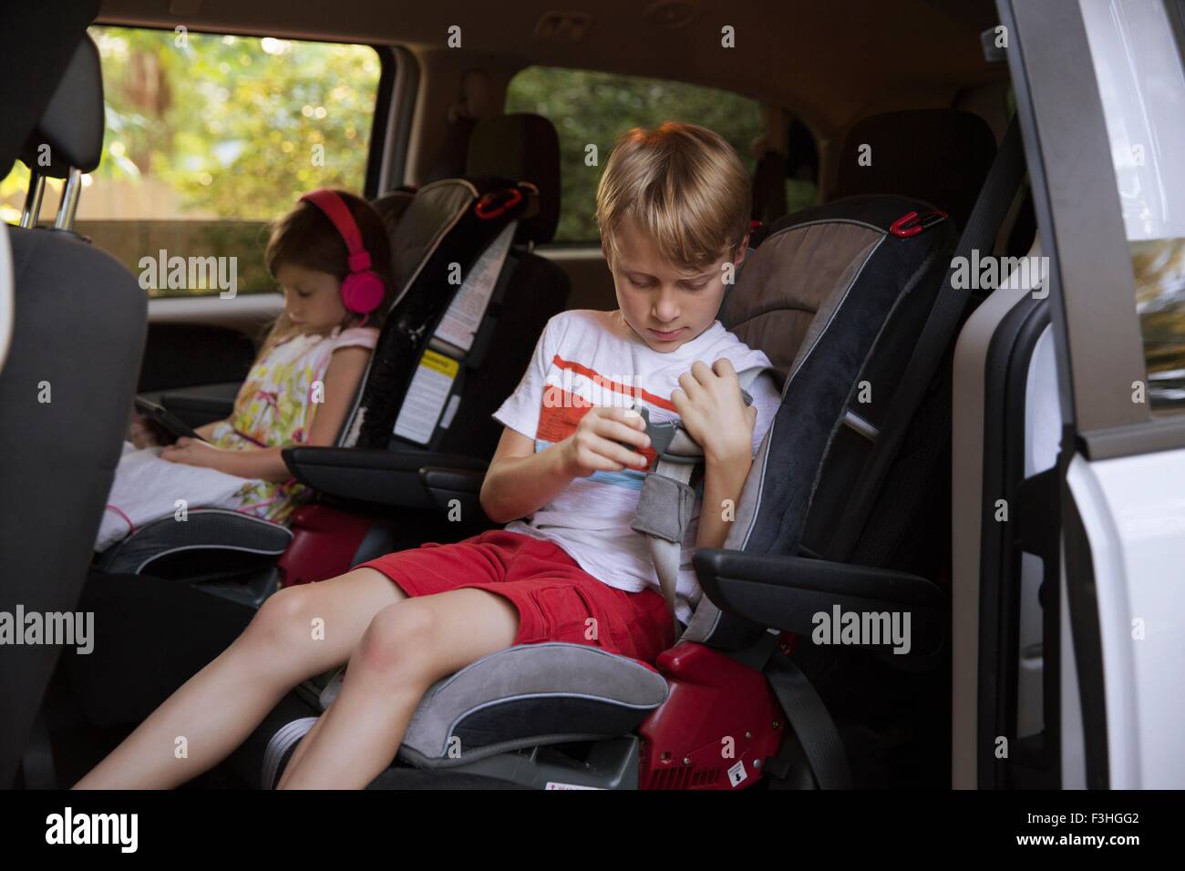 Girl using digital tablet whilst brother fastens seat belt in car back seat Stock Photo