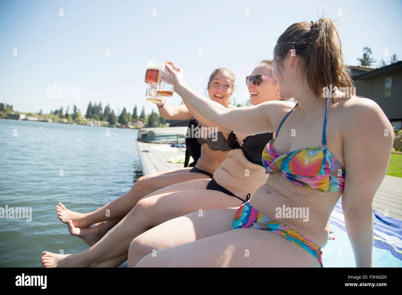 Young women wearing bikinis toasting with beer on waterfront pier, Lake Oswego, Oregon, USA Stock Photo