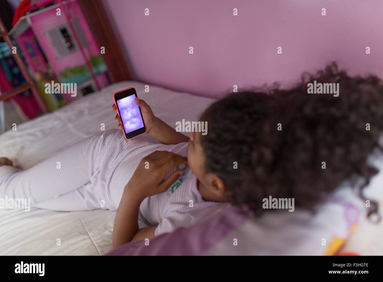 Over shoulder view of girl wearing pyjamas lying on bed looking at smartphone Stock Photo