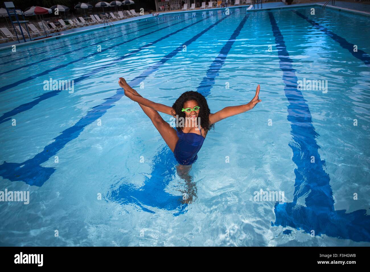Portrait of girl standing on one leg in swimming pool wearing goggles, leg raised, looking at camera, smiling Stock Photo