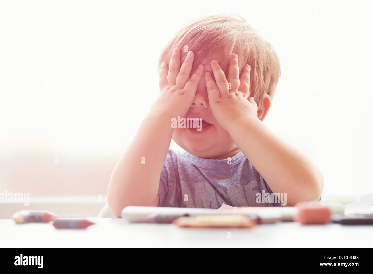 Boy sitting at table resting on elbows, mouth open, covering eyes with hands Stock Photo