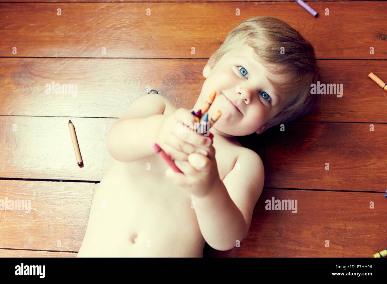 High angle view of boy lying on back on wooden floor holding crayons, looking at camera Stock Photo
