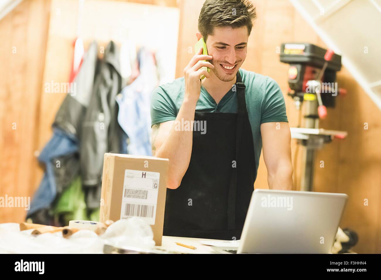 Young man in workshop standing at desk talking on telephone looking at laptop Stock Photo