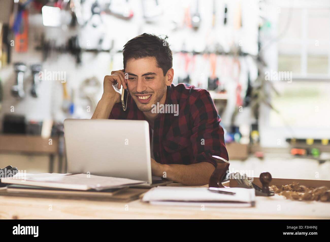 Young man sitting at desk in workshop talking on phone using laptop smiling Stock Photo