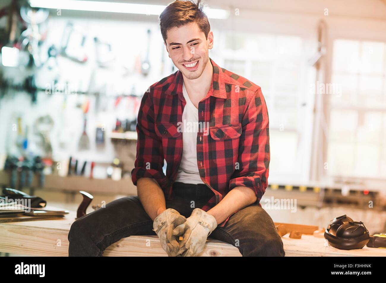Young man sitting on workbench wearing protective gloves, looking away smiling Stock Photo