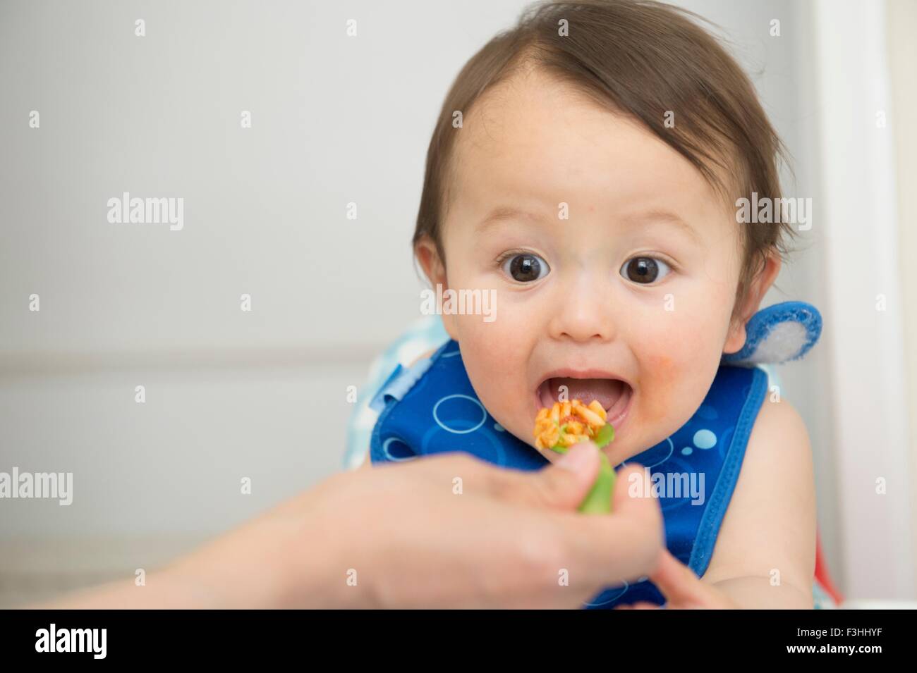 Hand of mother feeding baby food to open mouthed son in kitchen Stock Photo
