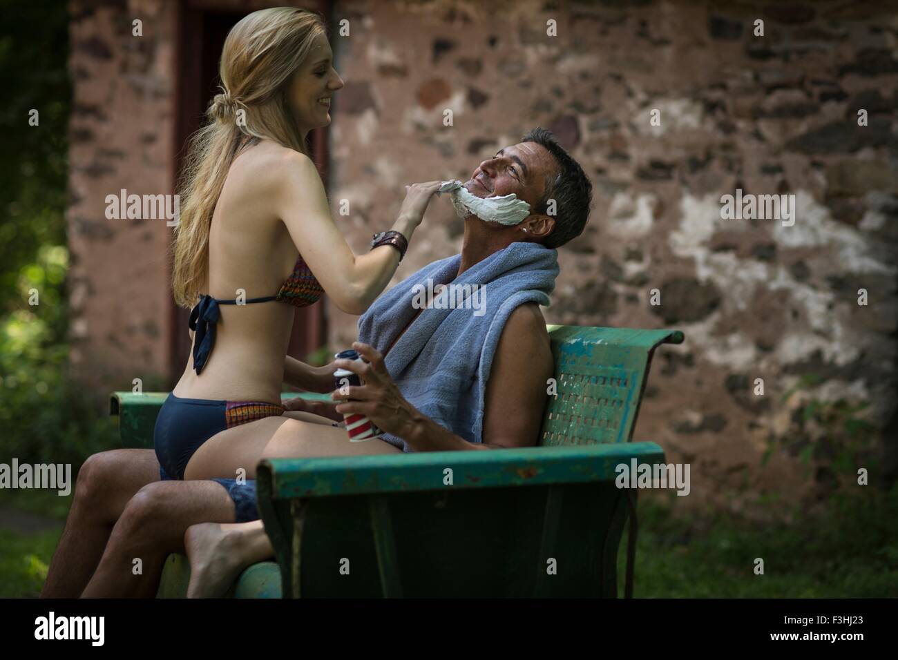 Young woman in bikini sitting on boyfriends lap applying shaving cream at holiday cottage Stock Photo