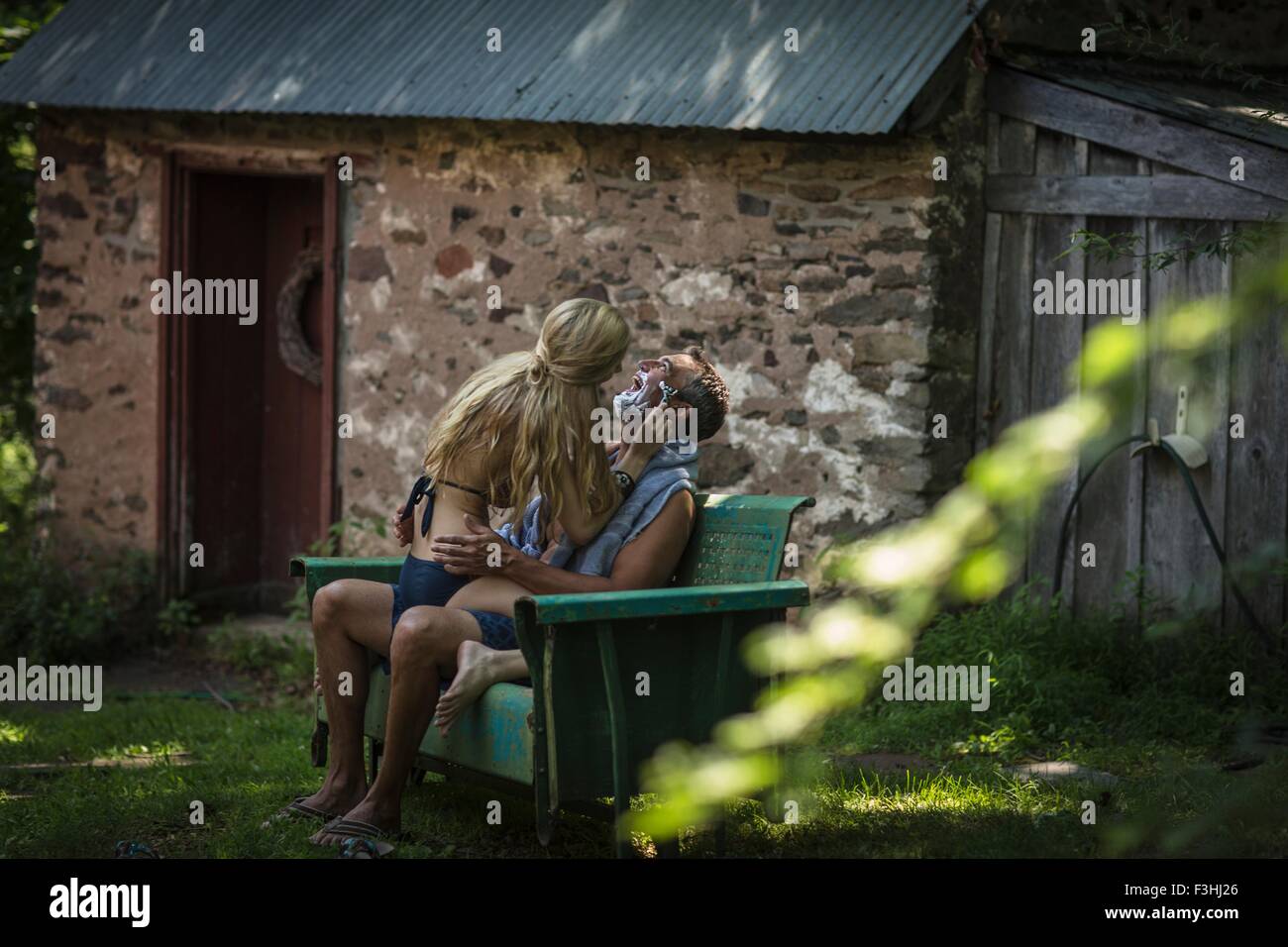 Young woman in bikini sitting on boyfriends lap shaving his chin at holiday cottage Stock Photo