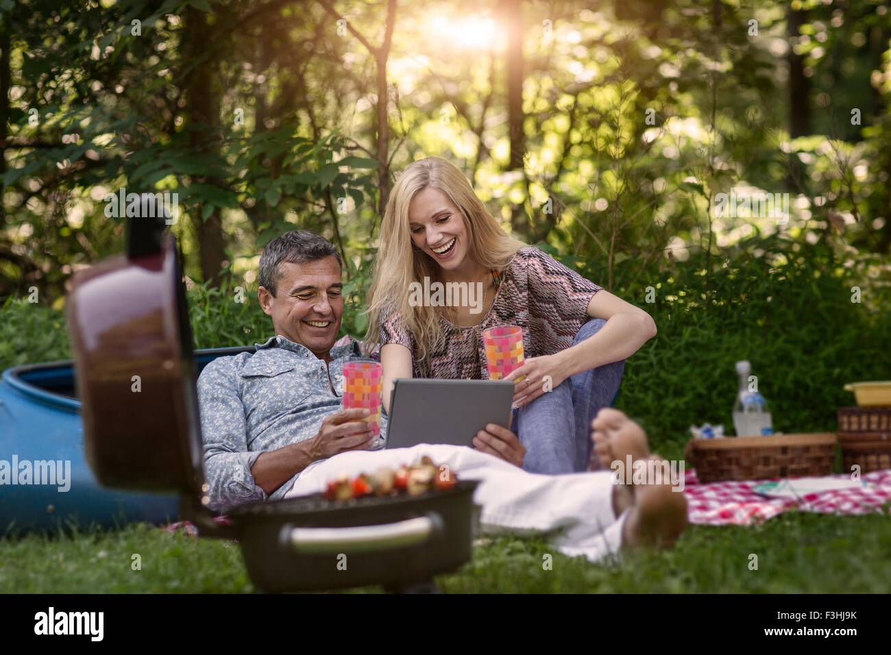 Mature man and girlfriend looking at digital tablet whilst having picnic barbecue Stock Photo