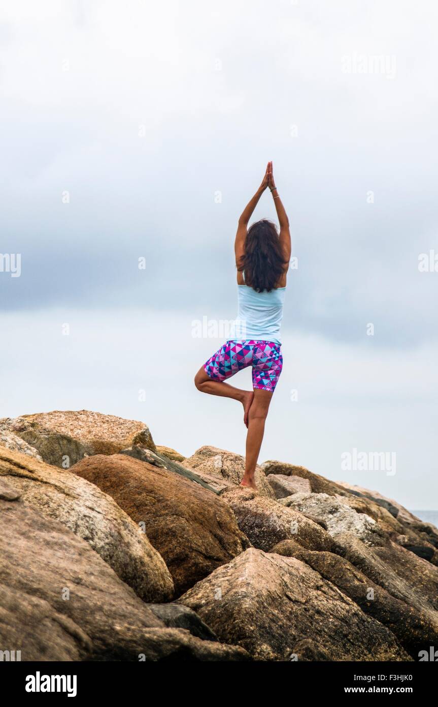 Rear view of mature woman practicing yoga tree pose on coastal rocks, Stock Photo