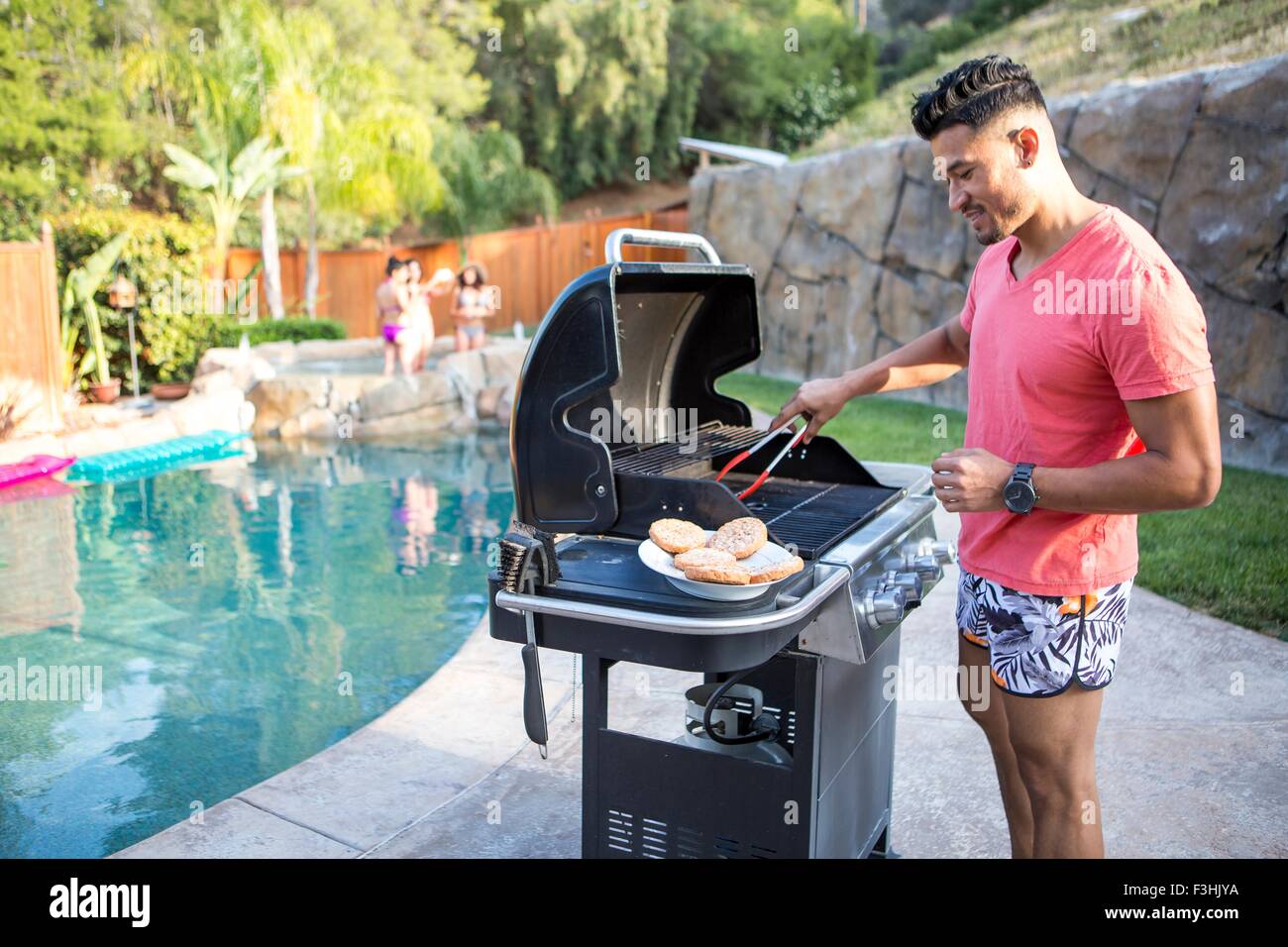 Young man cooking burgers on barbecue next to garden swimming pool Stock Photo