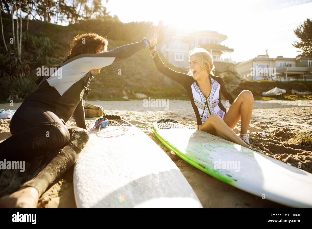 Couple sitting on beach, clapping hands in high five, surfboards beside them Stock Photo