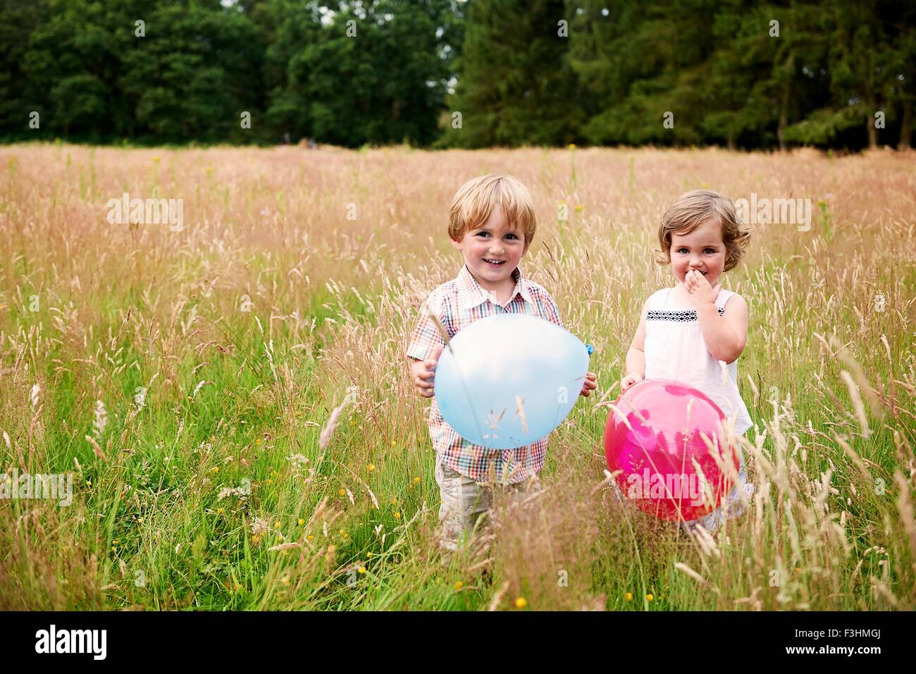 Brother and sister in tall grass holding balloon looking at camera smiling Stock Photo