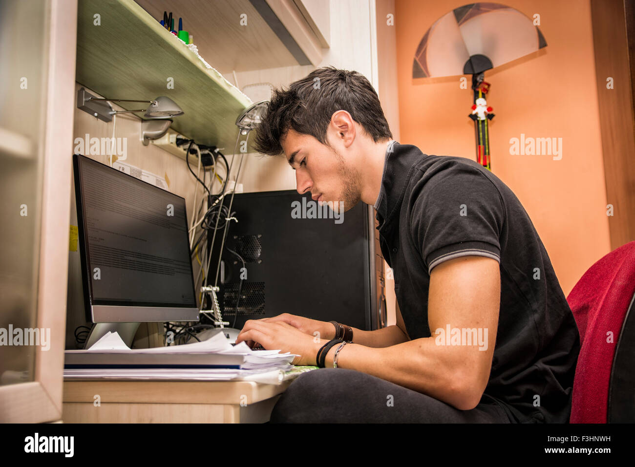 Waist Up Profile of Young Attractive Man with Dark Hair, Sitting at Computer Desk Working on Paper Homework or on His Start-up B Stock Photo