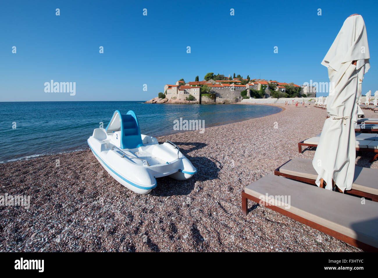 A catamaran and sun umbrellas on the beach in front of Sveti Stefan (Saint Stephen) islet and hotel resort, Montenegro Stock Photo