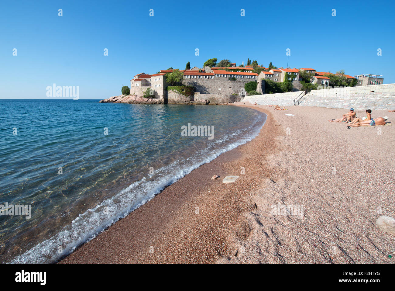 Tourists sun bathing on the beach in front of Sveti Stefan (Saint Stephen) islet and hotel resort, Montenegro Stock Photo
