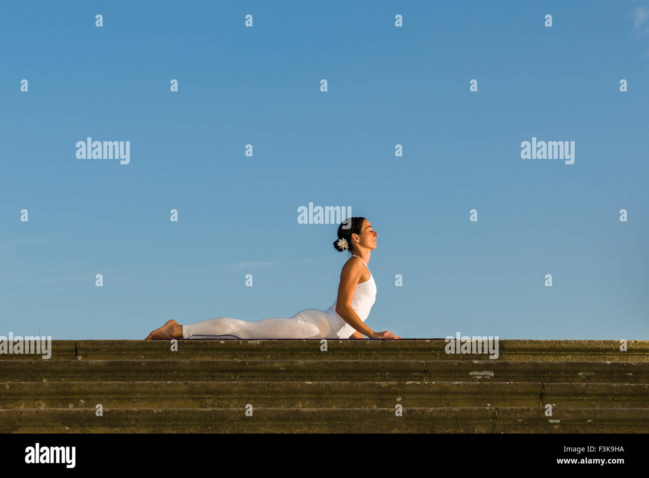 Young woman, wearing a white body suit, is practising Hatha-Yoga outdoor, showing the pose: bhujangasana, cobra pose Stock Photo