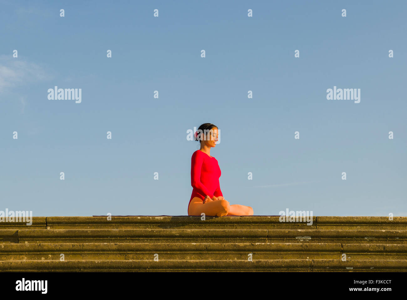 Young woman, wearing a red-orange body suit, is practising Hatha-Yoga outdoor, showing the pose: padmasana, lotus pose Stock Photo