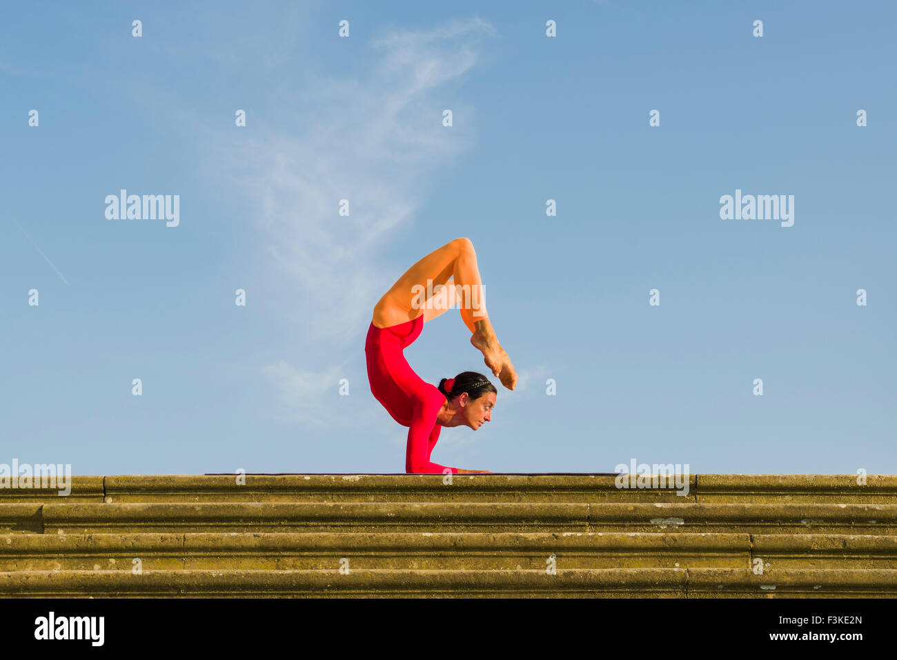 Young woman, wearing a red-orange body suit, is practising Hatha-Yoga outdoor, showing the pose: vrischikasana, scorpion pose Stock Photo