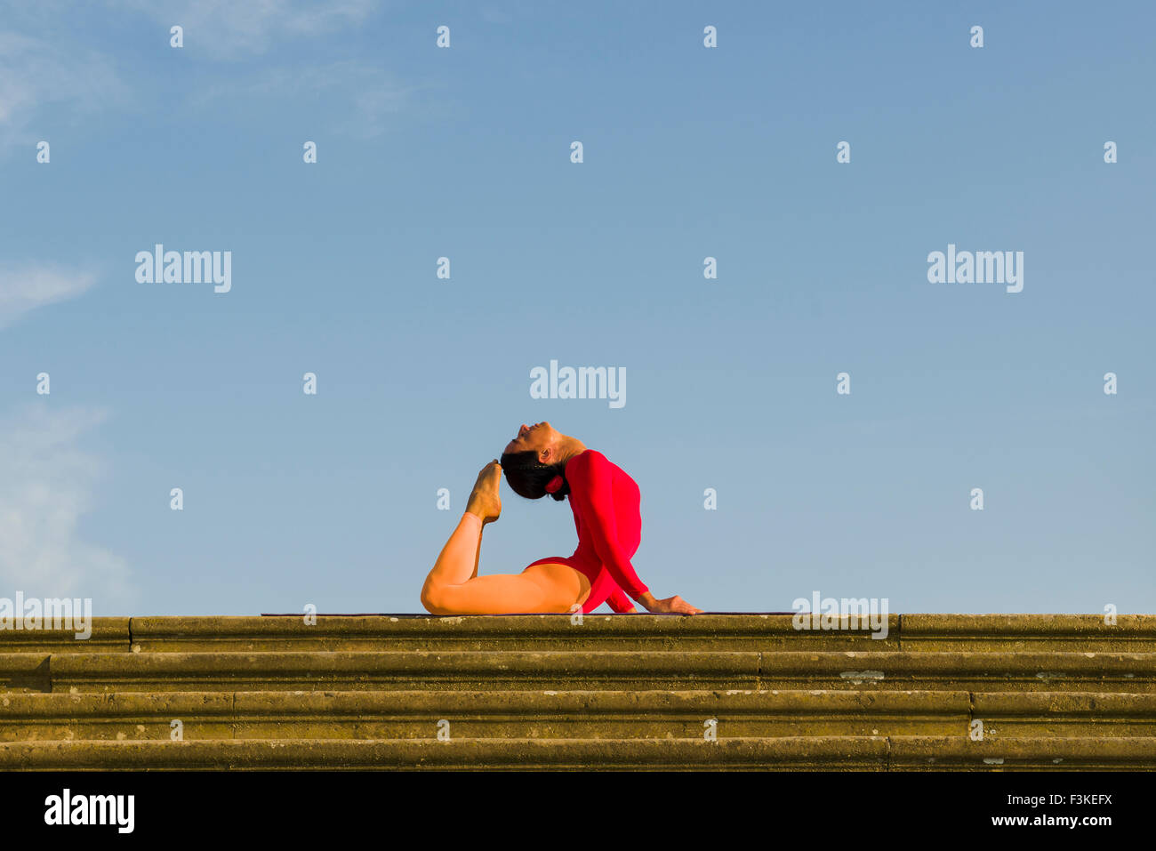 Young woman, wearing a red-orange body suit, is practising Hatha-Yoga outdoor, showing the pose: kapotasana, dove pose / pigeon  Stock Photo