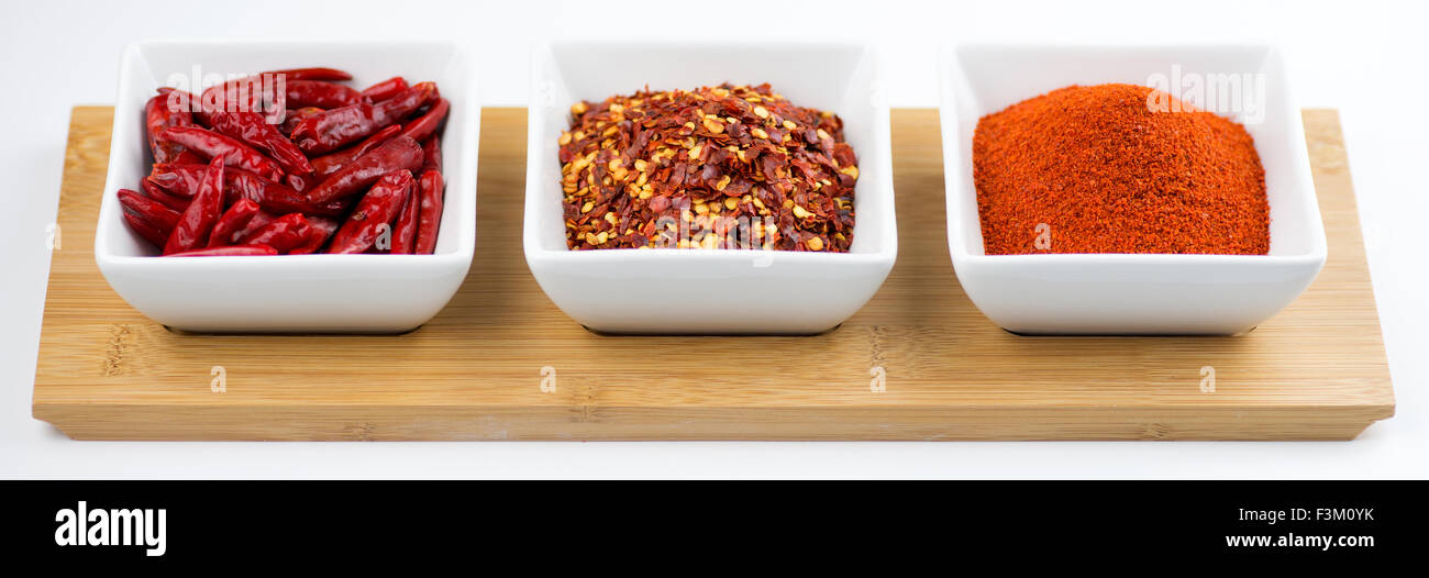 Chili peppers, flakes and cayenne powder in white bowls on a wooden tray against a white background Stock Photo