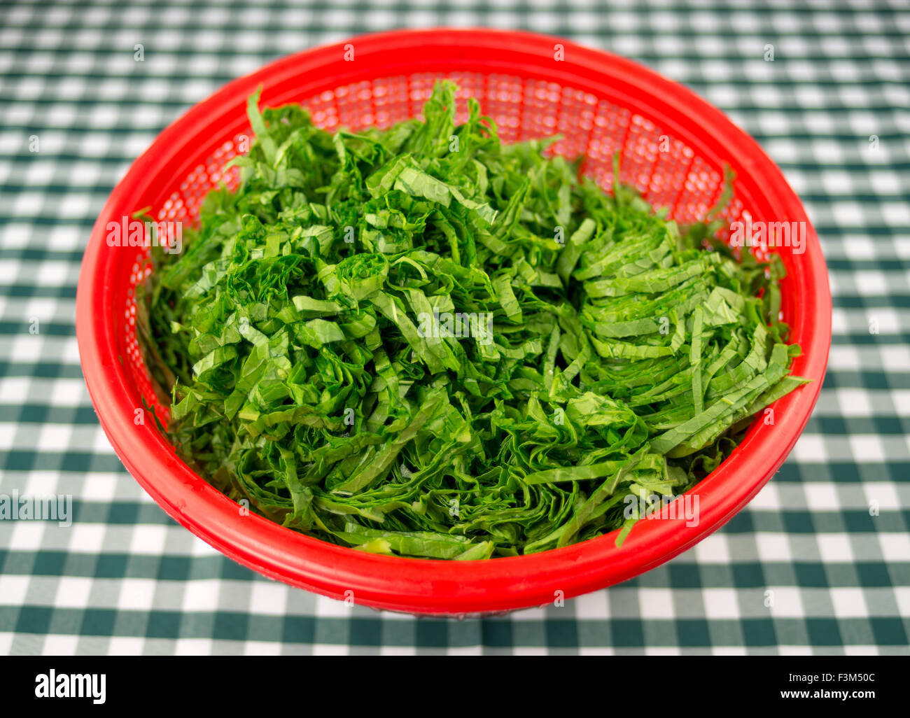 Freshly chopped raw mustard leaves in a red strainer bowl, against a green and white checkered tablecloth Stock Photo