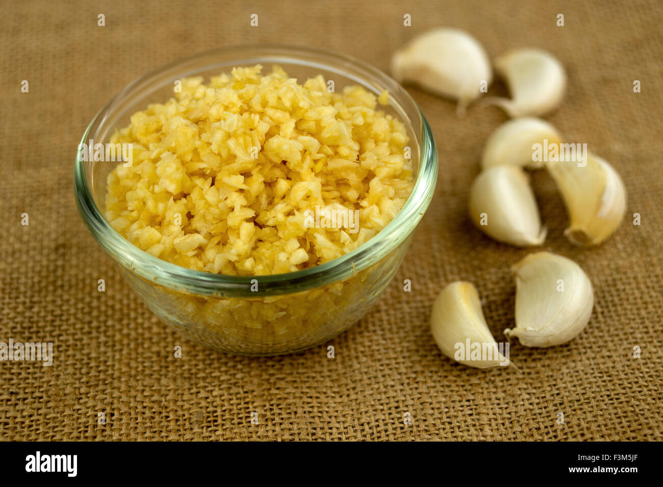Closeup of finely chopped garlic in a glass bowl with garlic cloves in the background against a sack cloth texture. Stock Photo