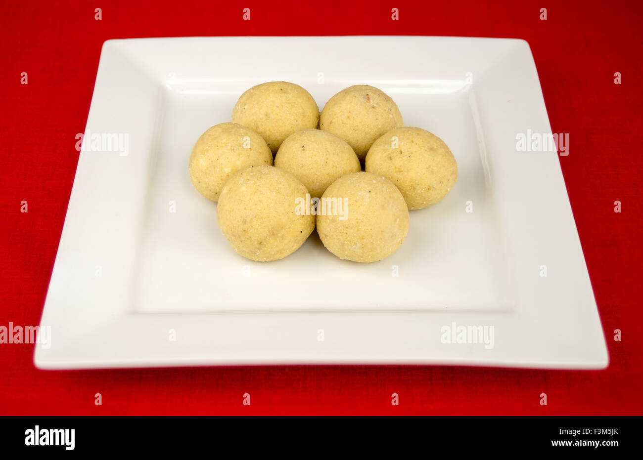 Ball shaped Indian laddu (laddoo) dessert plated on a square ceramic platter against a red tablecloth. Stock Photo