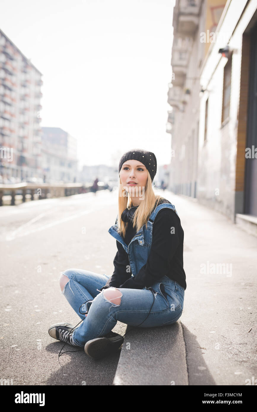 young beautiful blonde straight hair woman in the city, sitting on the  ground, overlooking right, holding a smartphone, pensive Stock Photo - Alamy