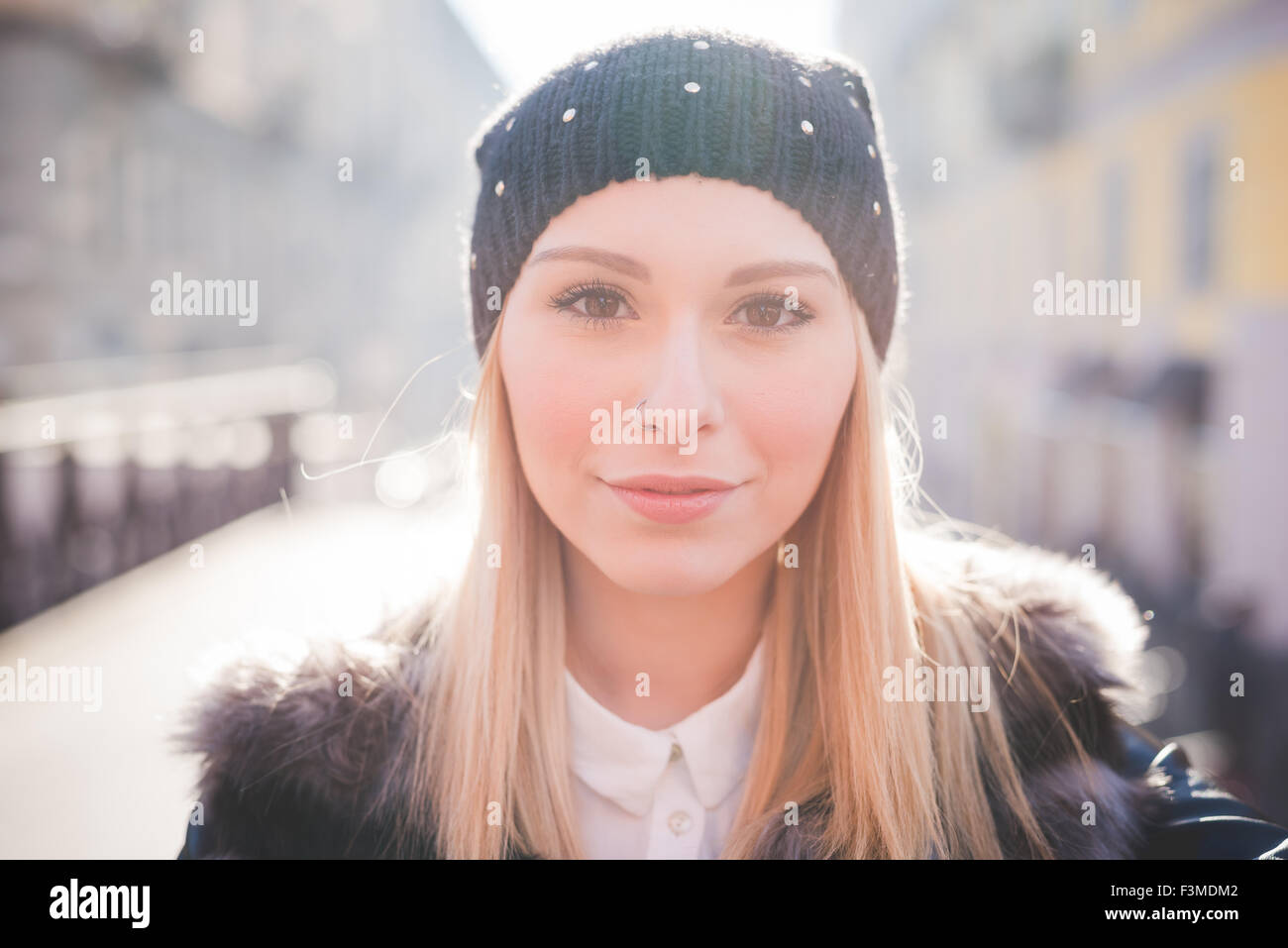 Portrait of young beautiful blonde straight hair woman in the city, looking in camera, smiling - wearing a black hat, a furry brown jacket - carefreeness, youth concept Stock Photo