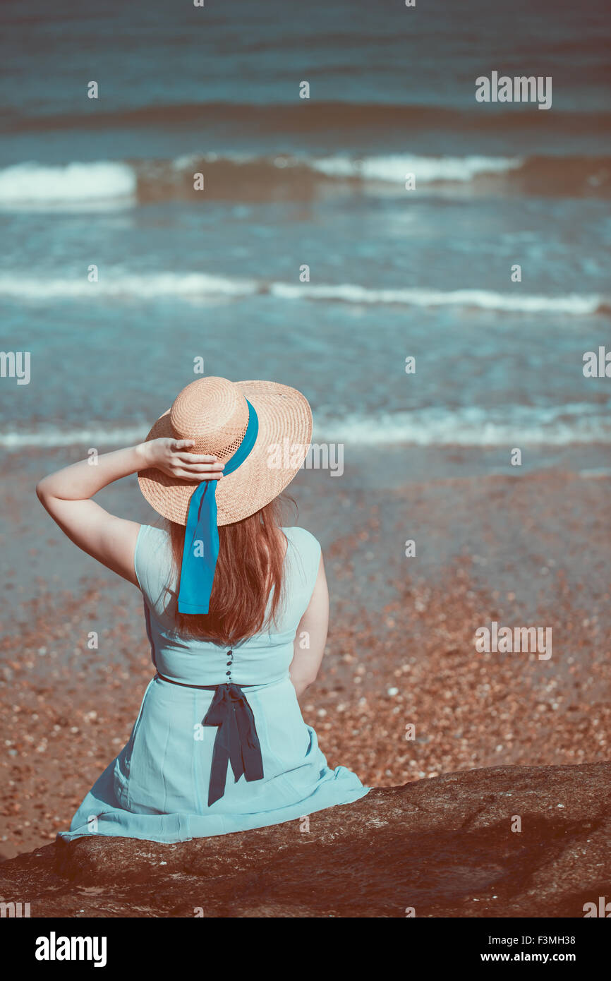 Beautiful young historical girl sitting on the beach in a blue dress and wearing a straw hat Stock Photo