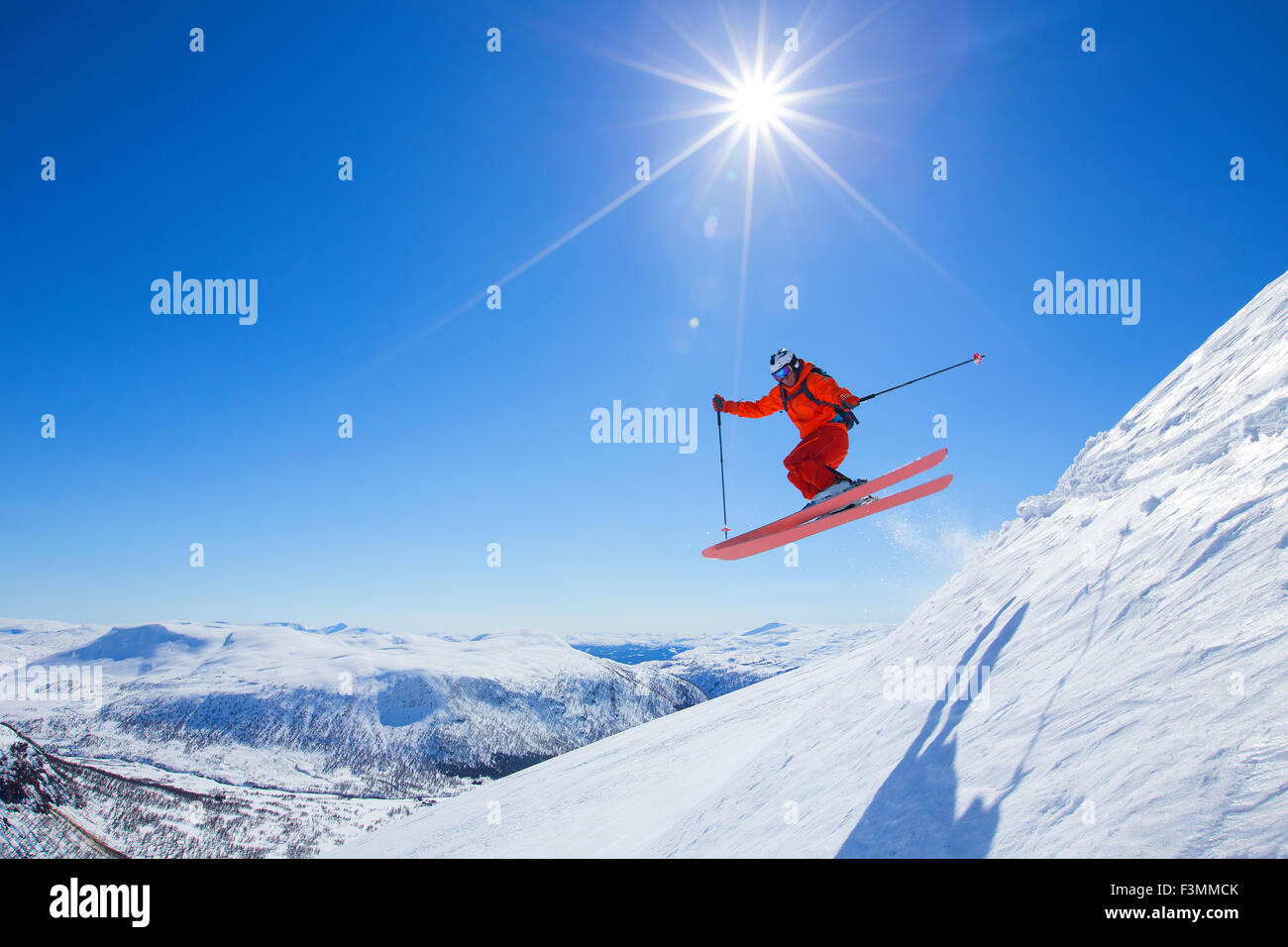 A male freerider in a red suit is jumping from a snow ridge. The sun is shining, the sky is blue. Stock Photo
