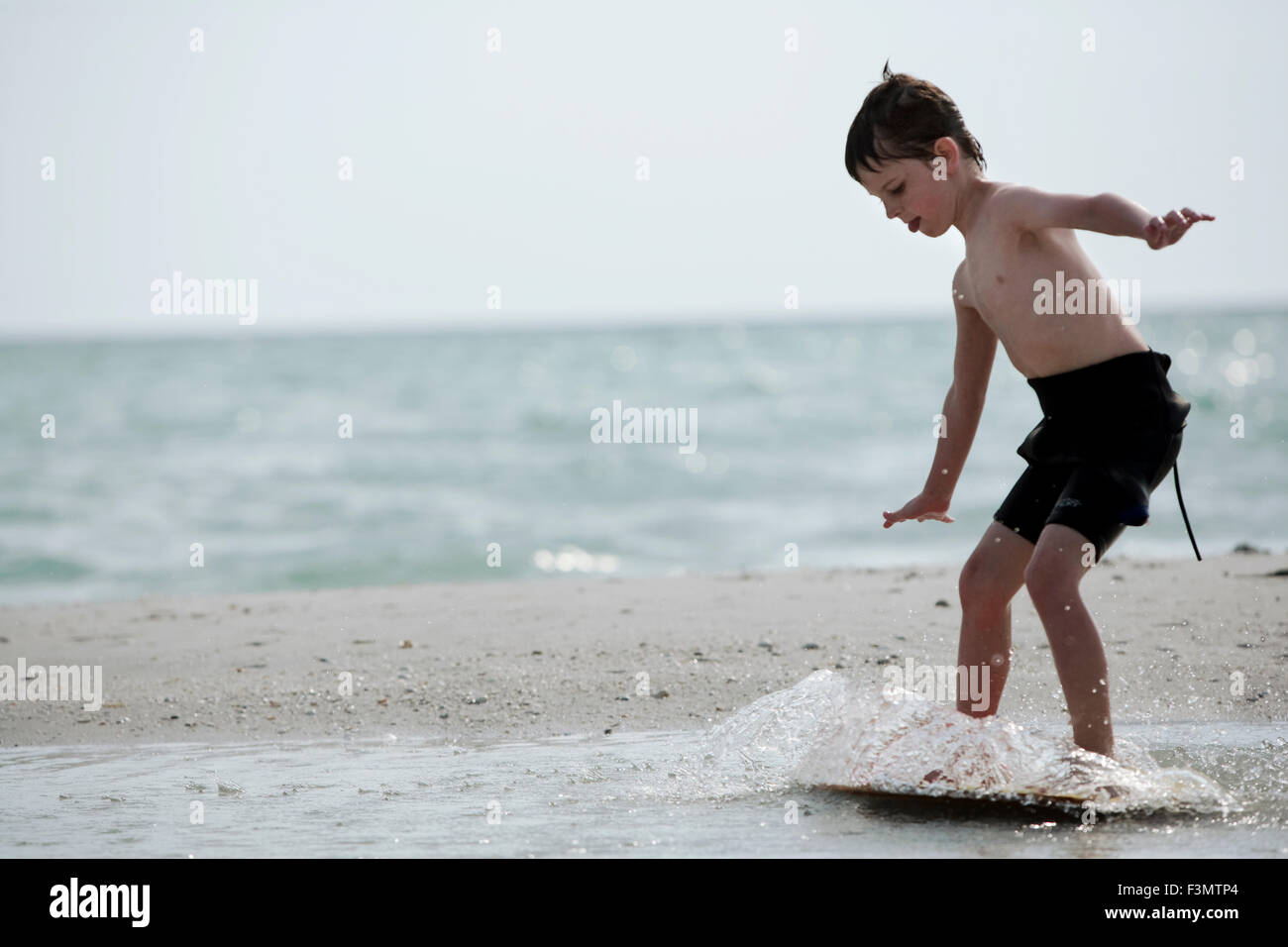 A young boy in a wetsuit surfs on his skim board at the shore. Stock Photo