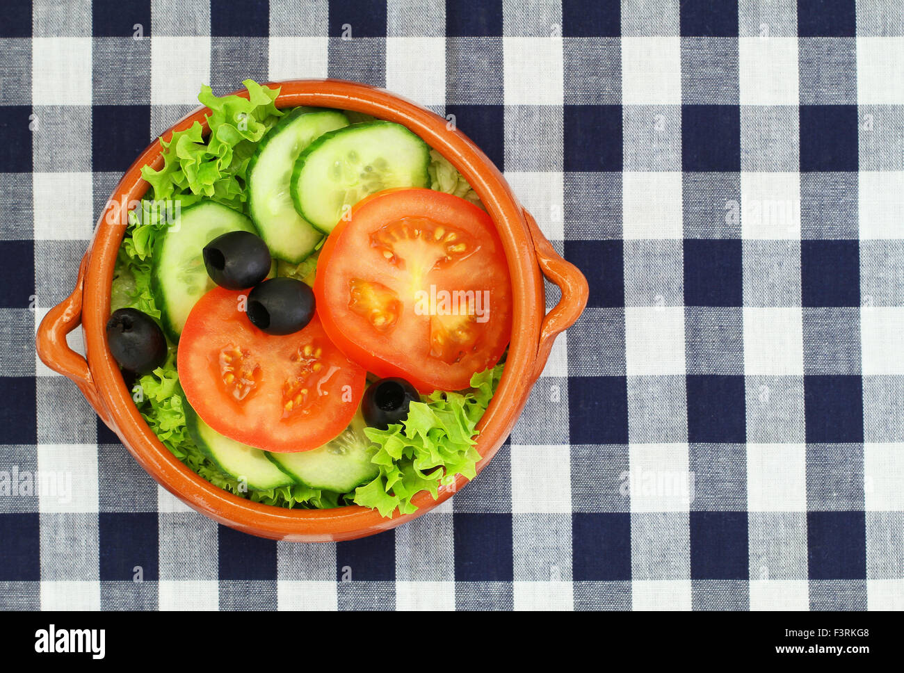 Green salad consisting of lettuce, tomatoes, cucumbers and black olives in traditional clay bowl on checkered cloth with copy sp Stock Photo