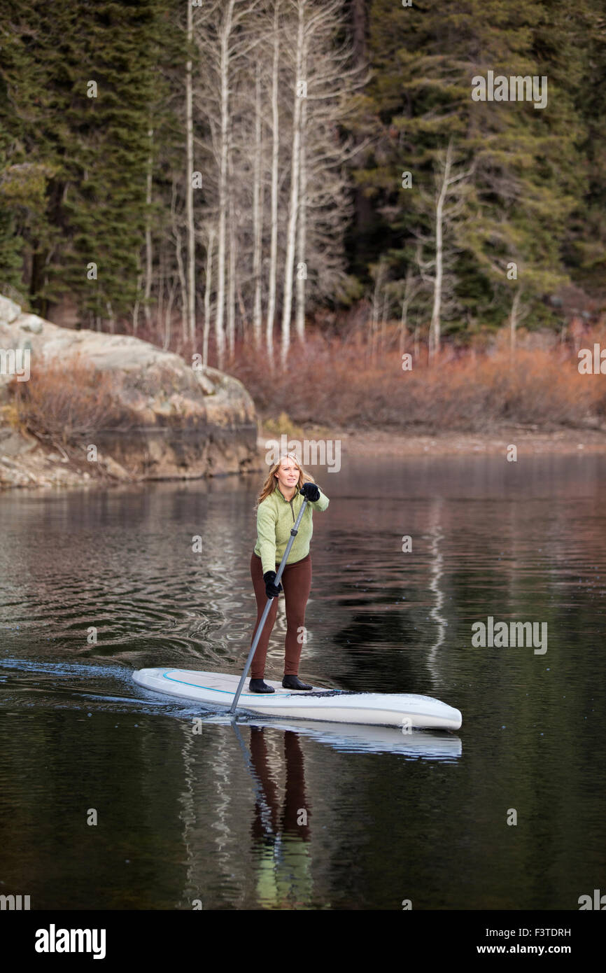 A young woman dressed for cold Fall weather paddling her paddle board on a small mountain lake in Northern California. Stock Photo