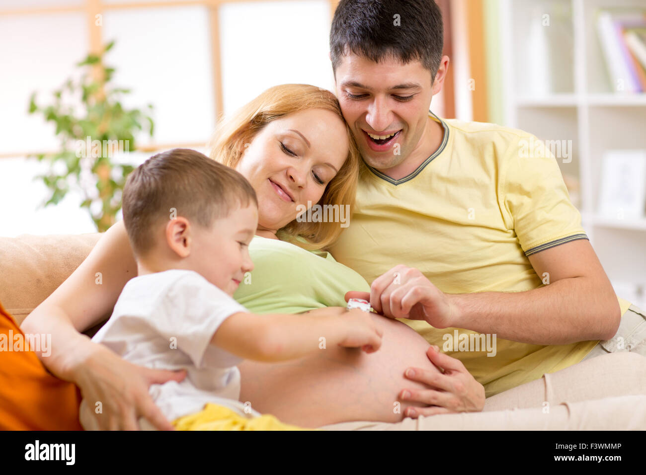 Father and his son have a fun playing with tiny car on wife belly. Pregnant woman with family relaxing on sofa in living room. Stock Photo
