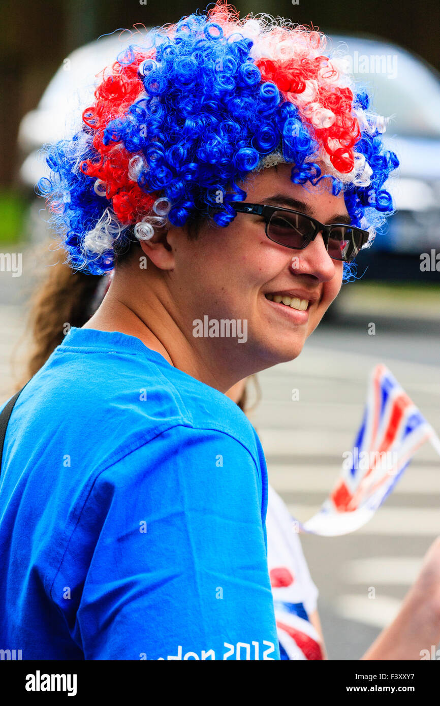 Man with red, white and blue hair, standing on side of street, wearing sunglasses and holding small Union jack flag. Turned towards viewer, smiling. Stock Photo