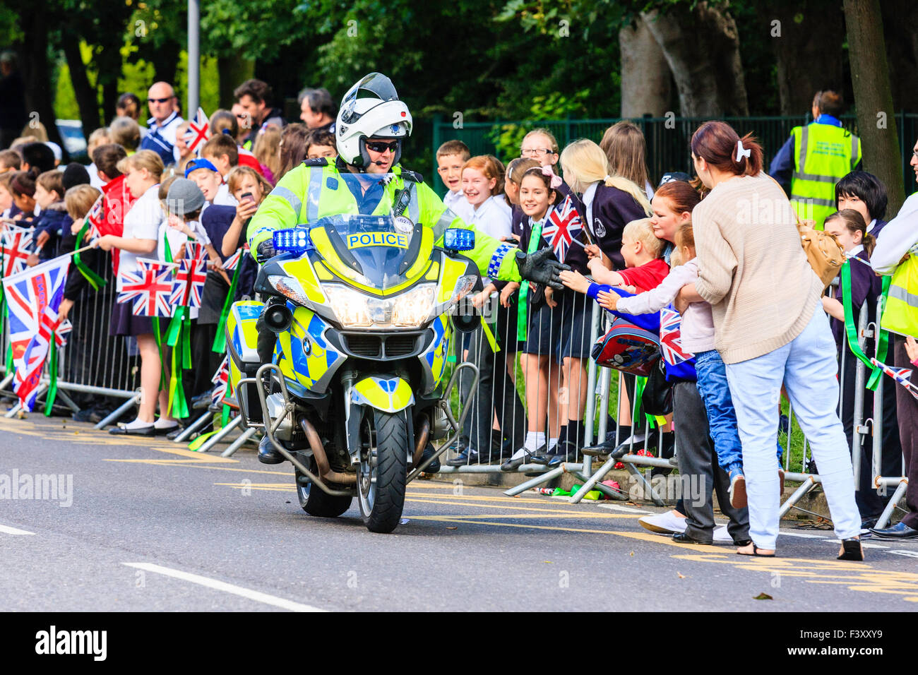 England. Main road with policeman on motorbike driving past cheering school children pushing against temporary railing lined with union jack flags. Stock Photo