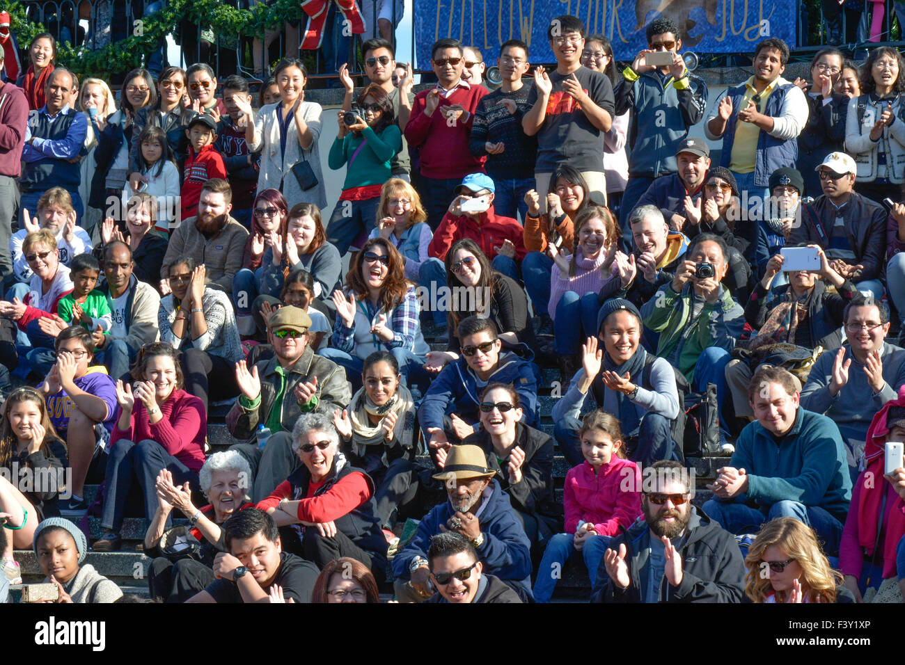 Crowd of ethnically diverse people of all ages, in the USA, some sitting,  some standing while laughing & applauding while watching performance art. Stock Photo