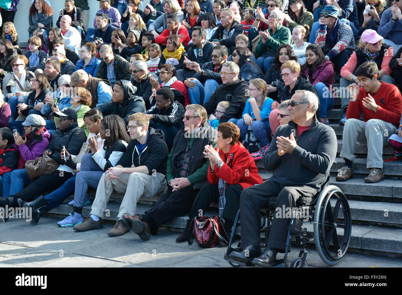 A large, diverse group of of multi-ethnic people sitting outside in rows as an audience watching street artists, with cheerful responses Stock Photo