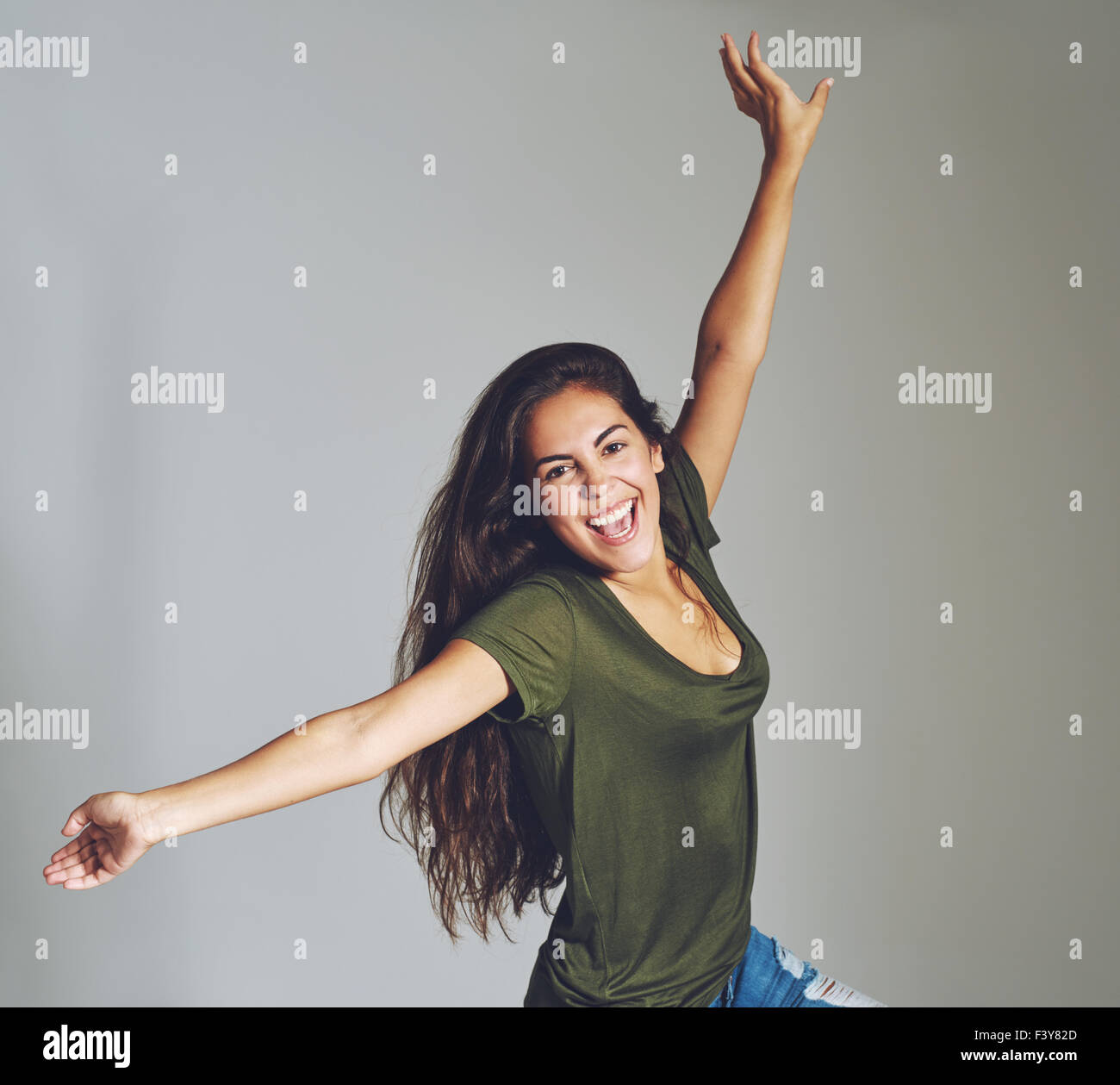 Vivacious attractive casual young woman celebrating throwing her arms in the air and laughing at the camera, over grey in square Stock Photo