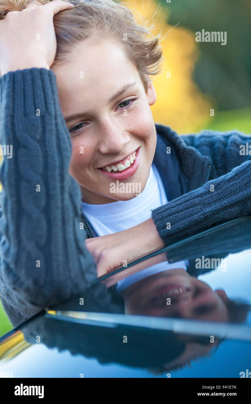 Young happy laughing male boy teenager blond child leaning on a car outside in summer sunshine Stock Photo