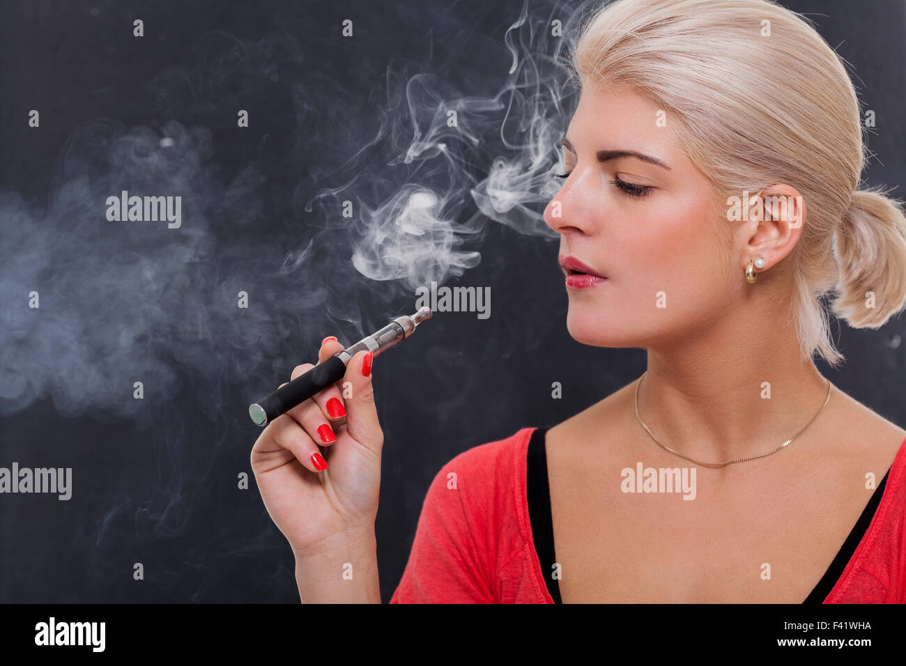 Stylish blond woman smoking an e-cigarette exhaling a cloud of smoke with her eyes closed in enjoyment, profile view on a dark background Stock Photo