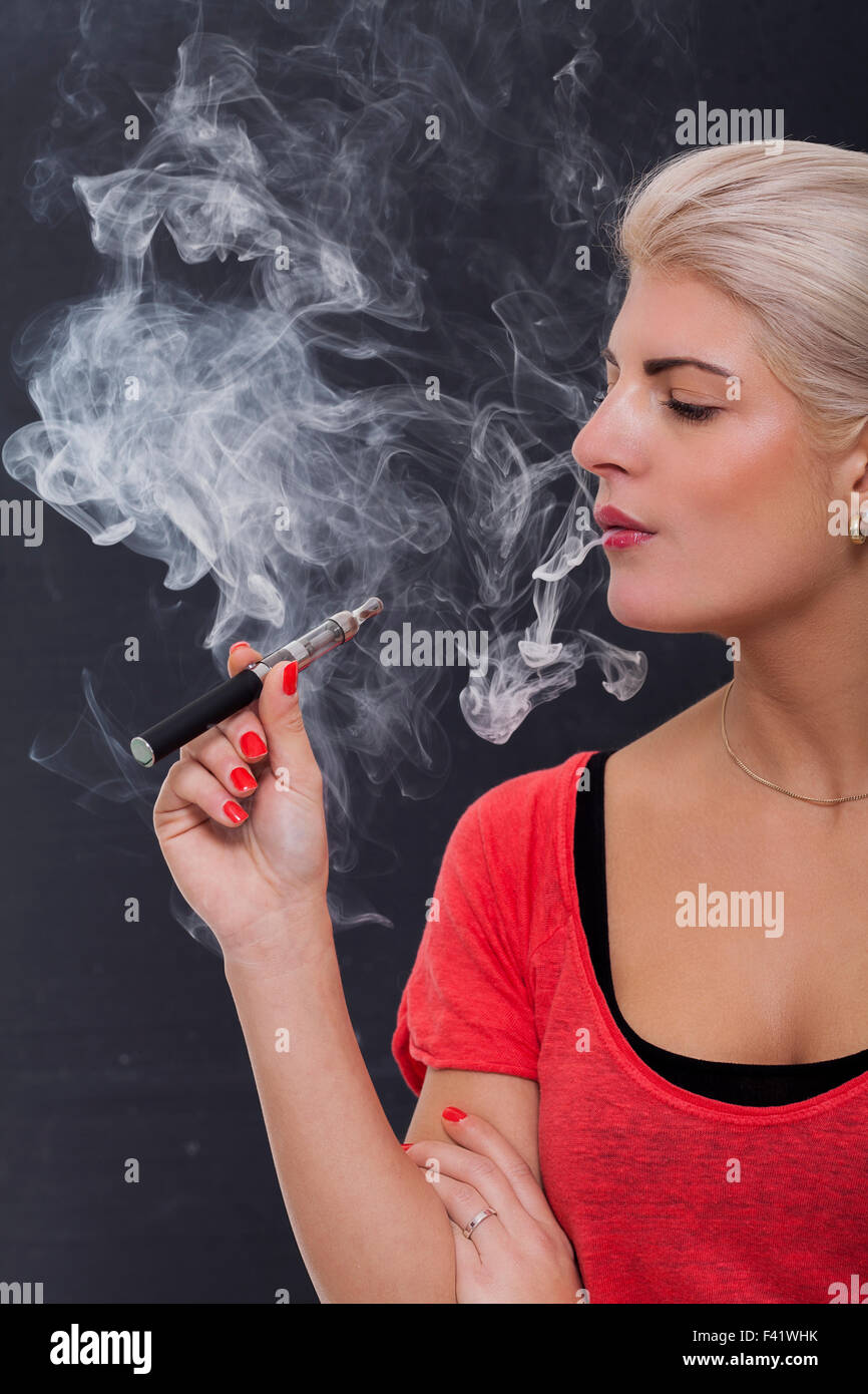 Stylish blond woman smoking an e-cigarette exhaling a cloud of smoke with her eyes closed in enjoyment, profile view on a dark b Stock Photo
