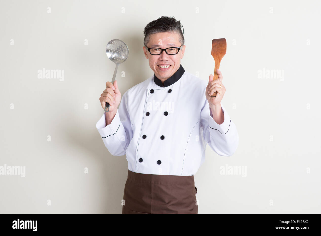 Portrait of confident 50s mature Asian male chef in uniform hands holding kitchen tools and smiling, standing on plain backgroun Stock Photo