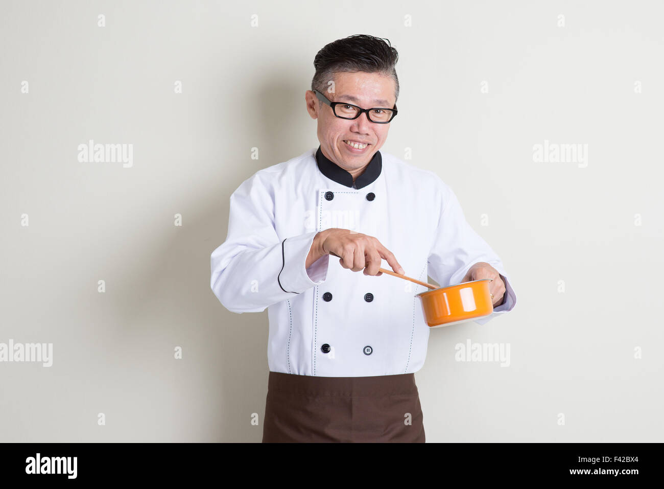 Portrait of mature Asian male chef in uniform cooking food, standing on plain background with shadow, copy space on side. Stock Photo