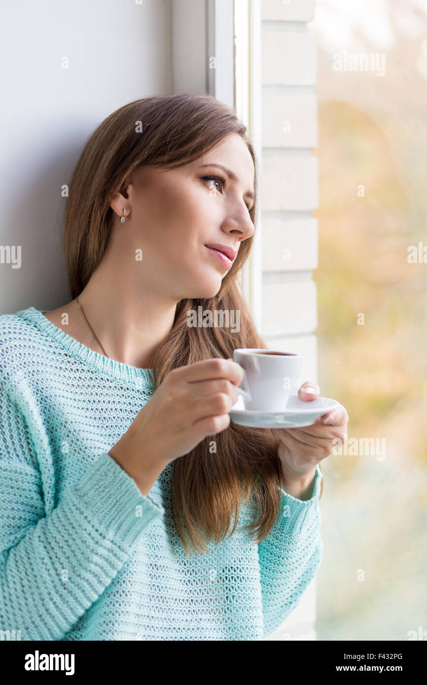 Young beautiful caucasian woman drinking her espresso at window sill. Gorgeous dreaming girl with cup of coffee Stock Photo
