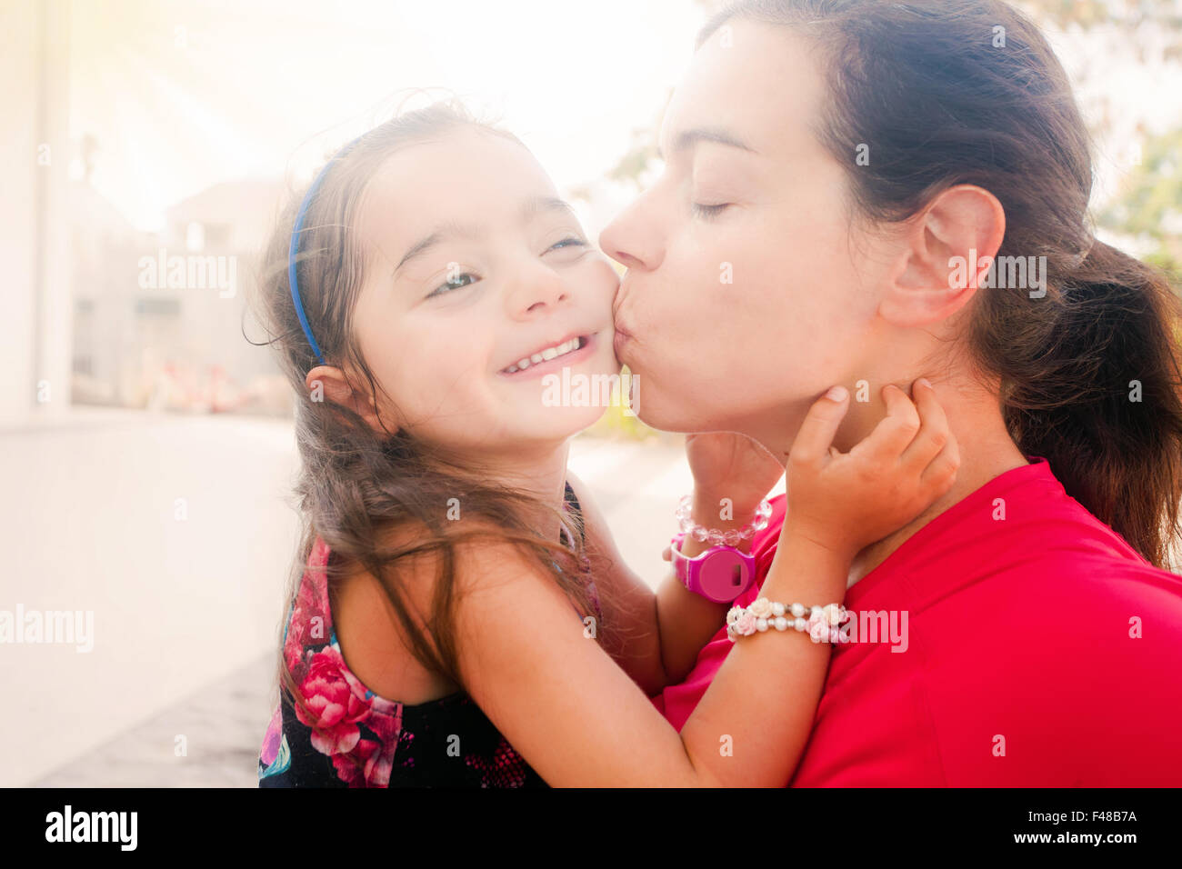 Young mother kissing her happy daughter on the cheek (sunset lighting, sun rays effect) Stock Photo