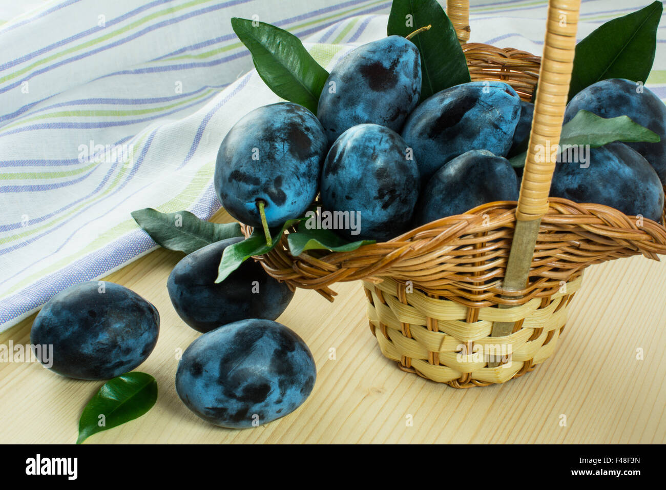 Ripe plums green leaves in small wicker basket on light wooden table with linen striped towel. Selective focus Stock Photo